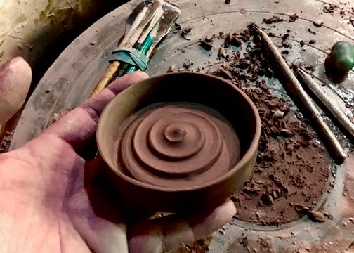 A hand holds a small clay bowl with a trimmed spiral pattern in its base, on a pottery wheel with trimming tools nearby